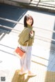 A woman in a green jacket and pleated skirt is standing on a sidewalk.